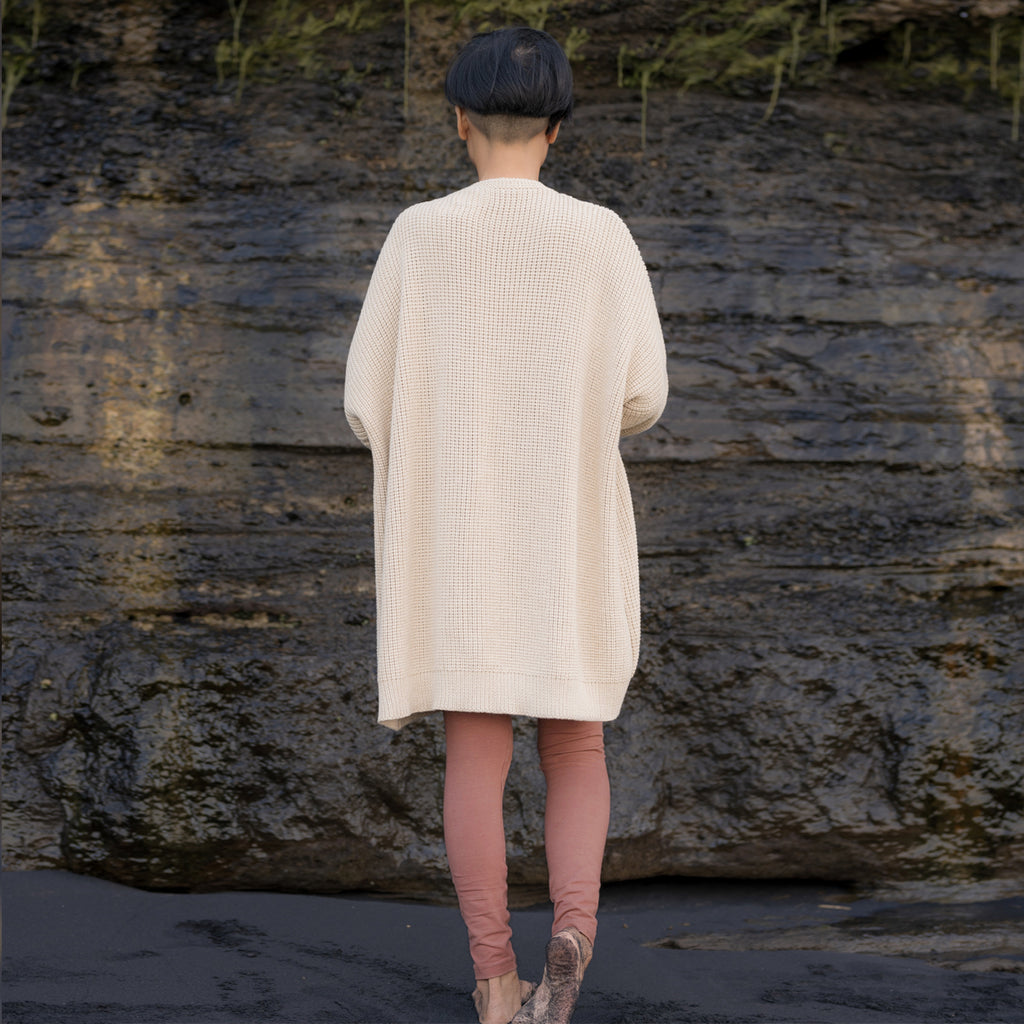 Sakti Rising Dhumavati Cardigan in oat color. Back view of a woman standing against a rock cliff, wearing an oversized cozy sweater.