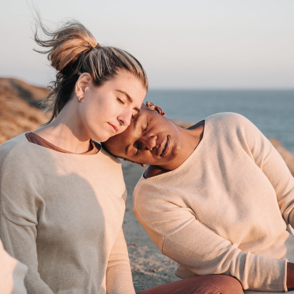 Caucasian woman and Black woman hugging on a sea cliff, wearing Sakti Rising Matangi Sweater in Oat. Ethical Sustainable yoga apparel.