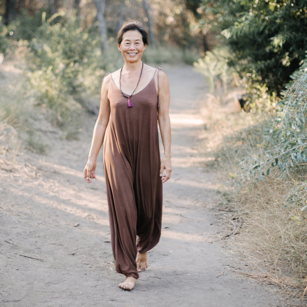 Asian woman walks along forest path smiling, wearing Sakti Rising Parvati Playsuit in Cacao. Ethical sustainable yoga apparel. 