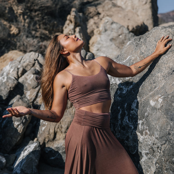 Asian woman stretches against a rock cliff, wearing Sakti Rising Yogini Bralette in Cacao. Sustainable, ethical yoga apparel. 