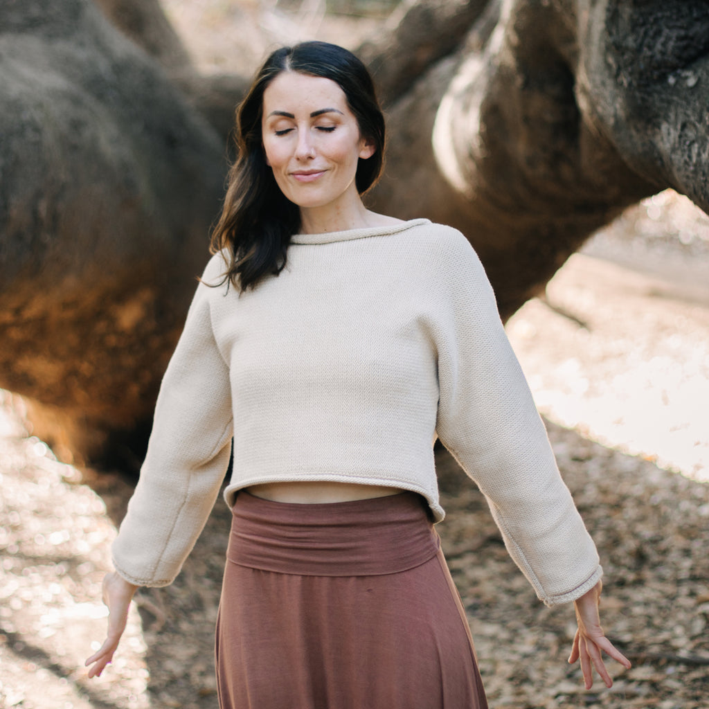 Woman standing with open palms by a tree. Wearing Sakti Rising Bagala Crop Sweater in Oat, sustainable and ethical yoga meditation apparel