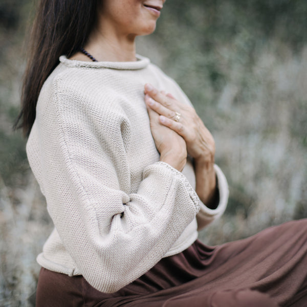 Asian woman seated with hands on heart. Wearing Sakti Rising Bagala Crop Sweater in Oat, sustainable and ethical yoga meditation apparel.