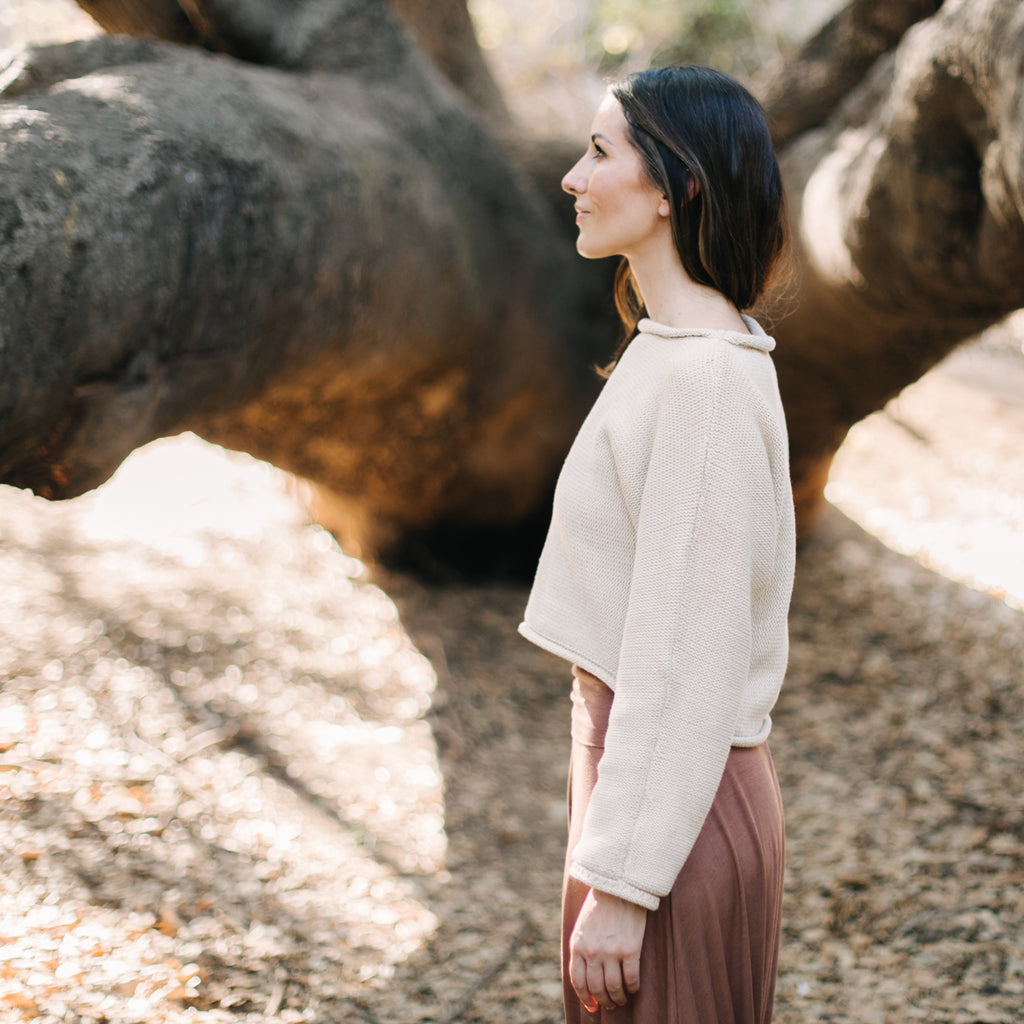 Caucasian woman standing in front of a tree, facing to the side. Wearing Sakti Rising Bagala Crop Sweater in Oat, sustainable and ethical yoga meditation apparel.
