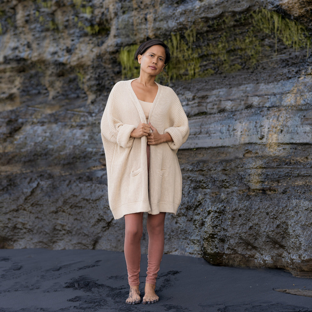 Asian woman stands in front of rock sea-cliff, wearing Sakti Rising Rudrani leggings in cacao, Yogini bralette, and Dhumavati cardigan. Sustainable, ethical yoga clothes.