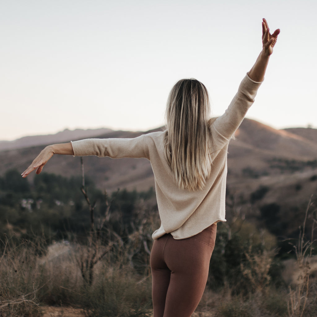 Woman on hilltop with arms raised, wearing Sakti Rising Matangi Sweater in Oat. Ethical Sustainable yoga apparel.
