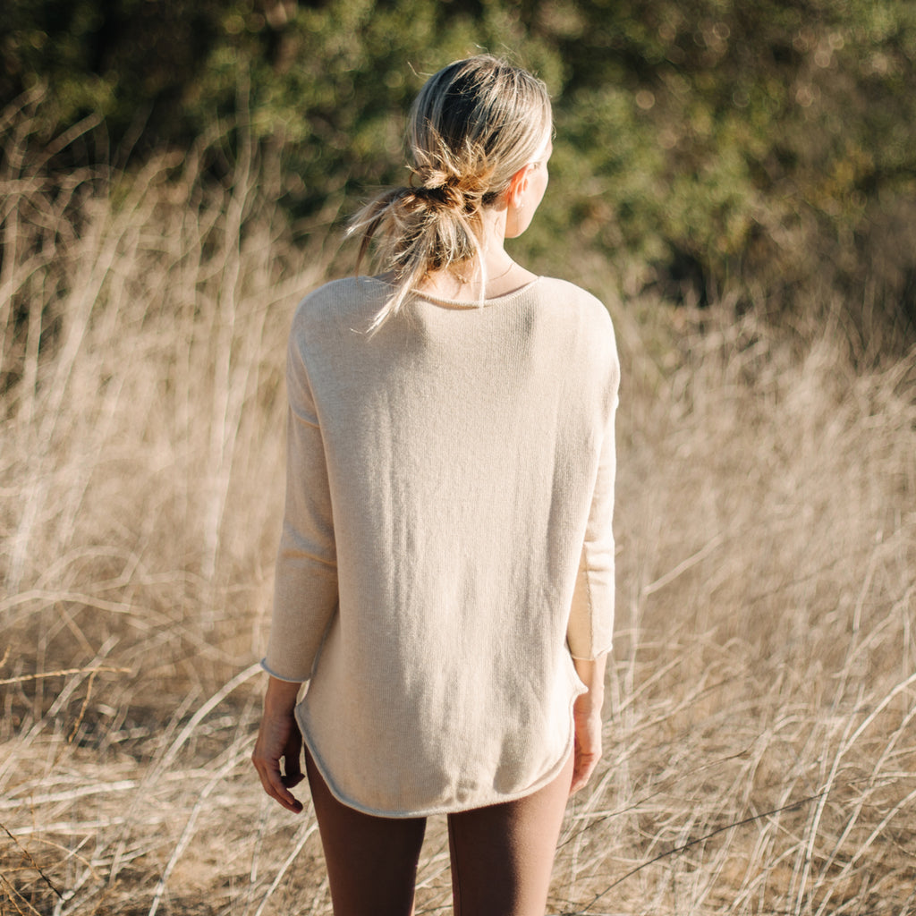 Back view of woman in field, wearing Sakti Rising Matangi Sweater in Oat. Ethical Sustainable yoga apparel.