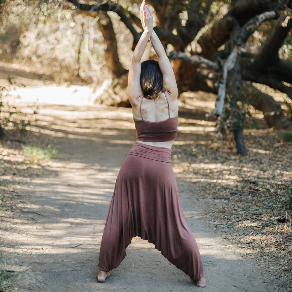 Back view of caucasian woman with arms raised in forest, wearing Sakti Rising Parvati Pants in Cacao. Ethical, Sustainable yoga apparel.
