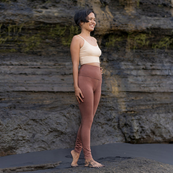 Asian woman stands in front of rock sea-cliff, wearing Sakti Rising Rudrani leggings in cacao and Yogini bralette in oat. Sustainable, ethical yoga clothes.