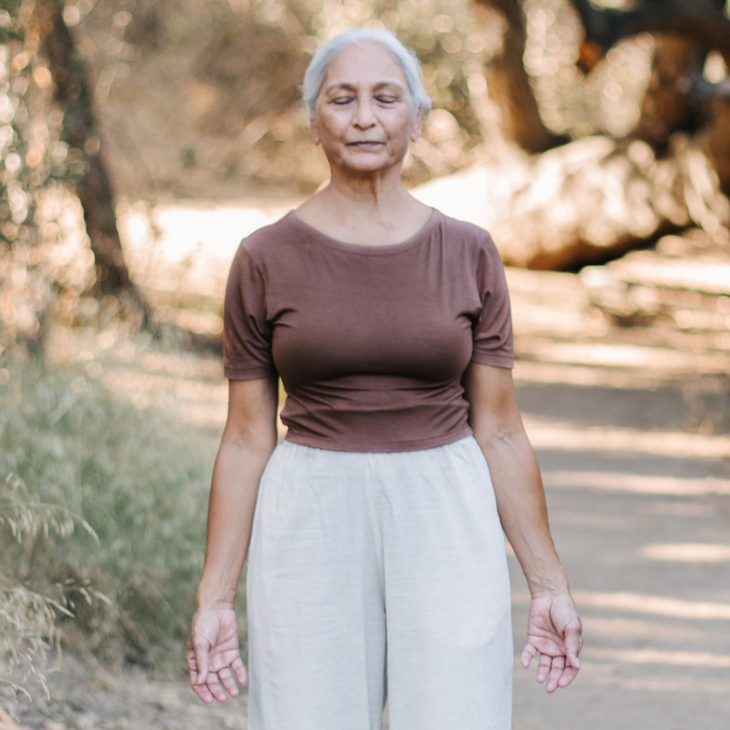 Elderly Indian woman stands with eyes closed in forest, wearing Sakti Rising Tara Tee in Cacao. Ethical sustainable yoga apparel.