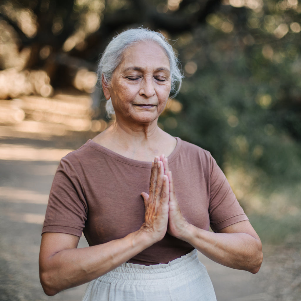 Elderly Indian woman praying, wearing Sakti Rising Tara Tee in Cacao. Ethical sustainable yoga apparel.